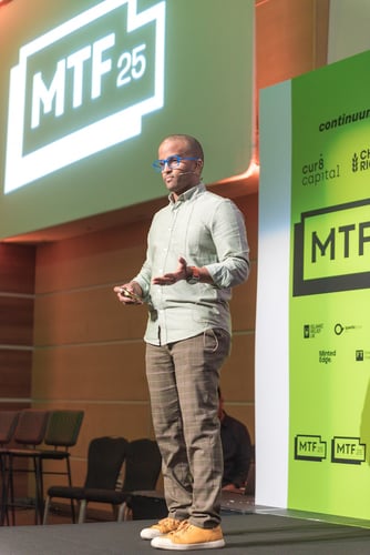 Ahmed standing at the stage of MTF 25 speaking in front of an audience, wearing green shirt, brown trousers, yellow shoes and blue glasses.