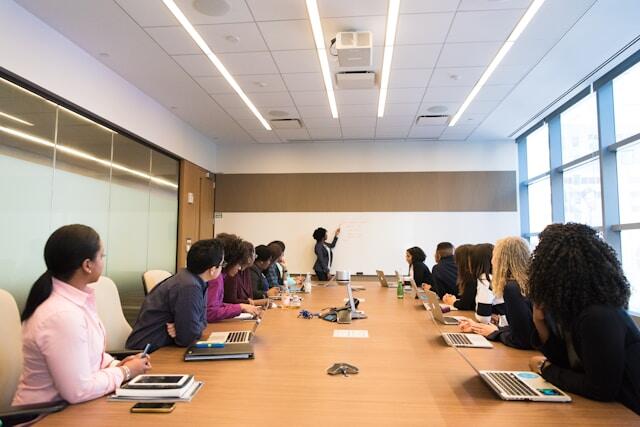 Board room with people sitting around a long table. One person is standing at one end doing a presentation on a whiteboard.